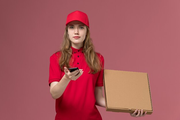 vrouwelijke koerier in rood uniform bedrijf levering voedseldoos en telefoon op lichtroze, uniform baan service werknemer levering meisje