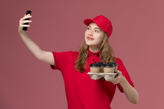 vrouwelijke koerier in rood uniform die bruine koffiekopjes houdt die selfie op roze, uniforme dienstbezorgerbaan nemen