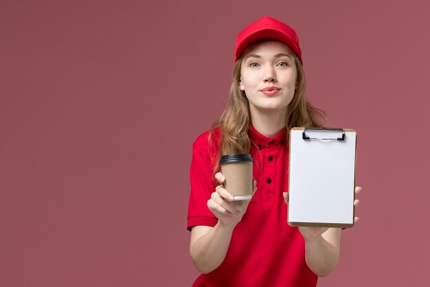 vrouwelijke koerier in rood uniform die koffiekop samen met blocnote op de lichtroze, baanuniforme levering van de werknemer houdt