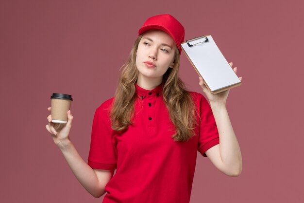 vrouwelijke koerier in rood uniform die koffiekop samen met blocnote op de lichtroze, baanuniforme levering van de werknemer houdt