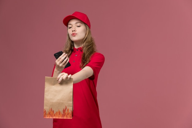 vrouwelijke koerier in rood uniform die voedselpakket houdt en haar telefoon gebruikt bij de levering van lichtroze, baanuniforme servicemedewerkers