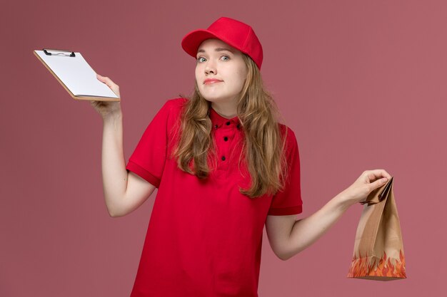 vrouwelijke koerier in rood uniform met bruin bezorgvoedselpakket en blocnote op de roze, uniforme dienstverlener