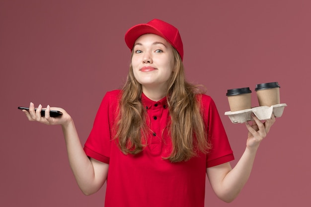 Vrouwelijke koerier in rood uniform met bruine bezorging koffiekopjes en telefoon op roze, uniforme dienstverlener