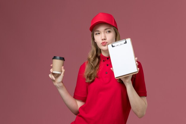 vrouwelijke koerier in rood uniform met koffie en blocnote op roze, uniforme dienstverlening