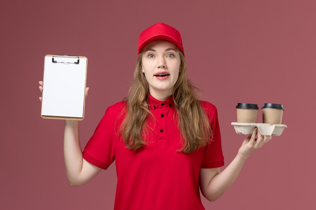 vrouwelijke koerier in rood uniform met koffiekopjes en blocnote op roze, uniforme dienstverlening