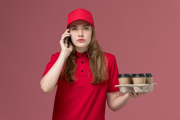 vrouwelijke koerier in rood uniform met koffiekopjes praten over de telefoon op lichtroze, baan uniforme werknemer dienstverlening