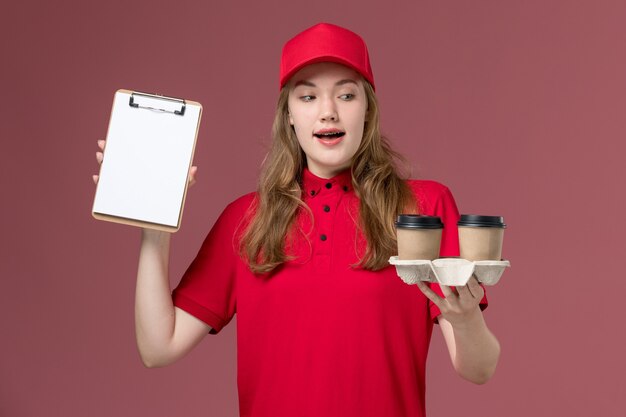 vrouwelijke koerier in rood uniform met koffiekopjes voor bezorging en blocnote op lichtroze, uniforme bezorger