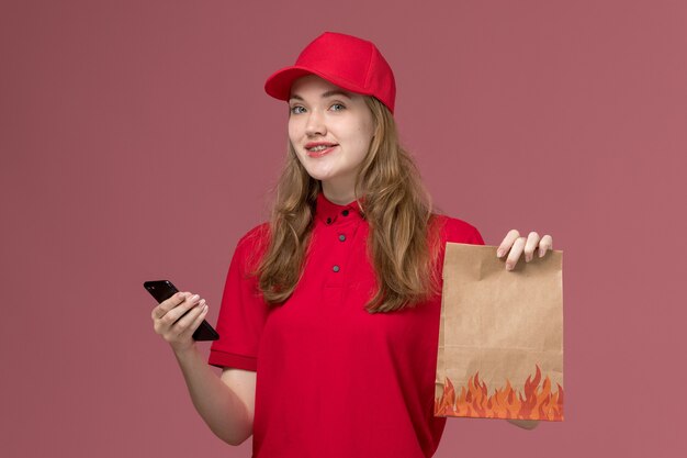 vrouwelijke koerier in rood uniform met papieren voedselpakket met telefoon op de lichtroze, baanuniforme levering van de werknemer