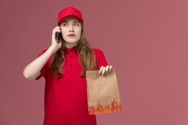 vrouwelijke koerier in rood uniform met voedselpakket praten aan de telefoon op roze, uniforme dienst levering baan werknemer