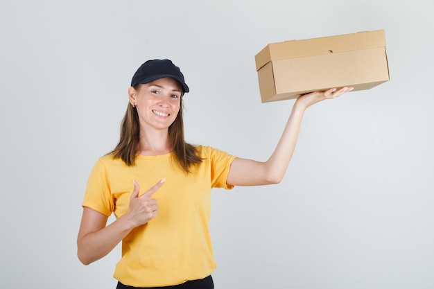 Vrouwelijke koerier wijzende vinger op kartonnen doos in t-shirt, broek, pet en op zoek blij