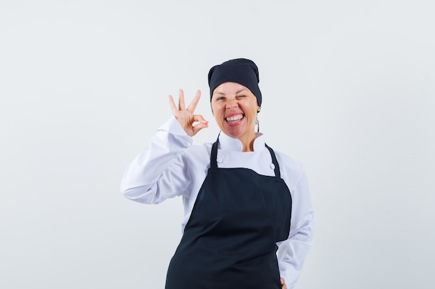 Vrouwelijke kok die ok gebaar in uniform, schort toont en zalig kijkt. vooraanzicht.