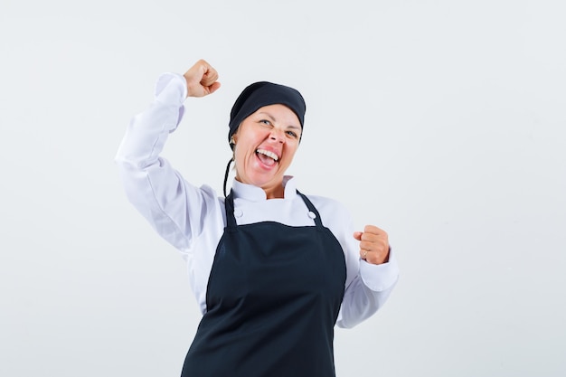 Vrouwelijke kok die winnaargebaar in uniform, schort toont en gelukkig kijkt. vooraanzicht.