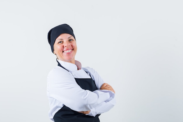 Vrouwelijke kok die zich met gekruiste armen in uniform, schort bevindt en gelukkig, vooraanzicht kijkt.