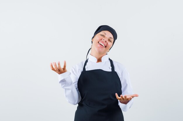 Vrouwelijke kok schalen gebaar maken in uniform, schort en vrolijk kijken. vooraanzicht.