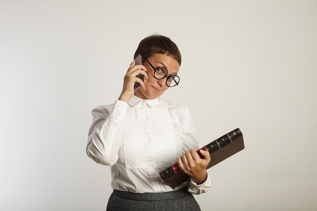 Vrouwelijke leraar in conservatieve outfit en ronde zwarte bril kijkt speels tijdens het gesprek aan de telefoon op de witte muur