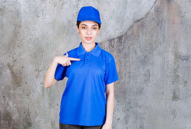 Vrouwelijke serviceagent in blauw uniform wijzend op zichzelf.