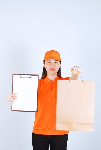 Vrouwelijke servicemedewerker in oranje dresscode die een kartonnen boodschappentas vasthoudt en de handtekeninglijst aan de klant presenteert