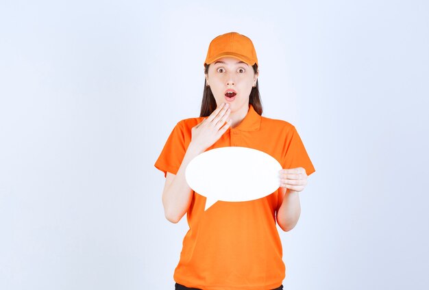 Vrouwelijke servicemedewerker in oranje dresscode met een ovaal infobord en kijkt verrast en doodsbang.