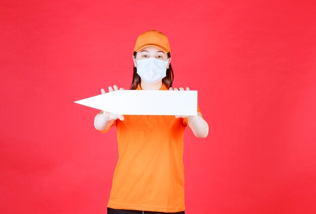 Vrouwelijke servicemedewerker in oranje kleur dresscode en masker met een pijl naar links.