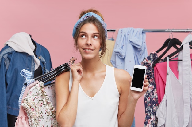 Vrouwelijke shopaholic die in boetiek staat, veel hangers met kleren meeneemt om te passen, met dromerige uitdrukking opzij kijkt, besluit wat te nemen, terwijl ze de moderne mobiele telefoon in de andere hand houdt