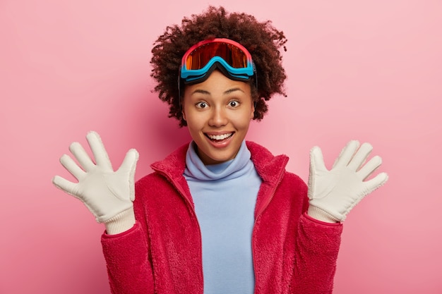 Vrouwelijke skiër met donkere huid heft palmen op in witte handschoenen, heeft plezier na het snowboarden, lacht vrolijk, draagt beschermend masker, coltrui en rode jas, heeft wintervakantie, geïsoleerd op roze achtergrond