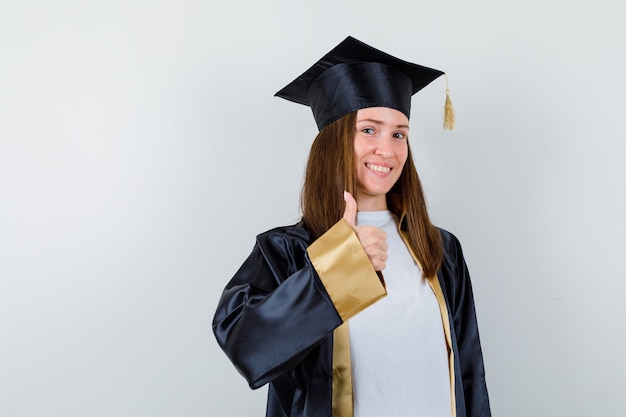 Vrouwelijke student die duim in graduatietoga toont en vrolijk kijkt. vooraanzicht.