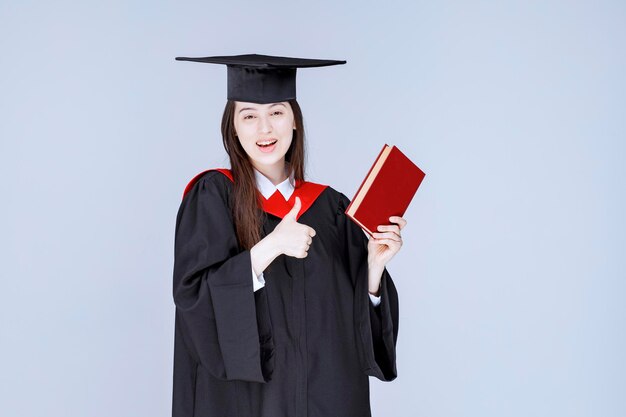 Vrouwelijke student die in academische toga rood boek houdt. Hoge kwaliteit foto
