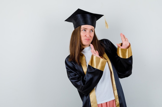 Vrouwelijke student in toga, vrijetijdskleding die stopgebaar toont en bang, vooraanzicht kijkt.