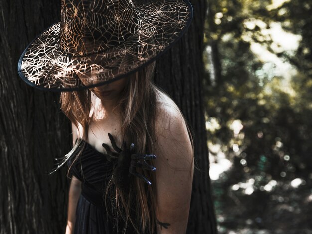 Vrouwelijke tovenares in hoed en spin op borst in zonovergoten struikgewas