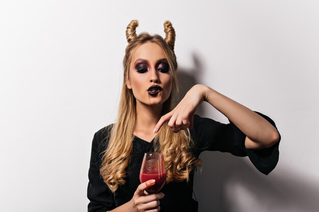 Vrouwelijke vampier die bloed drinkt uit wijnglas. Mooie blonde heks die van poition in halloween geniet.