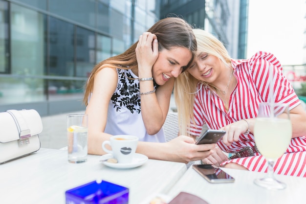 Vrouwelijke vrienden die bij koffie met koffie zitten die mobiele telefoon met behulp van