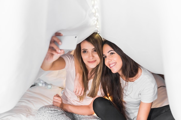 Gratis foto vrouwelijke vrienden die op bed zitten die selfie op smartphone onder het gordijn nemen