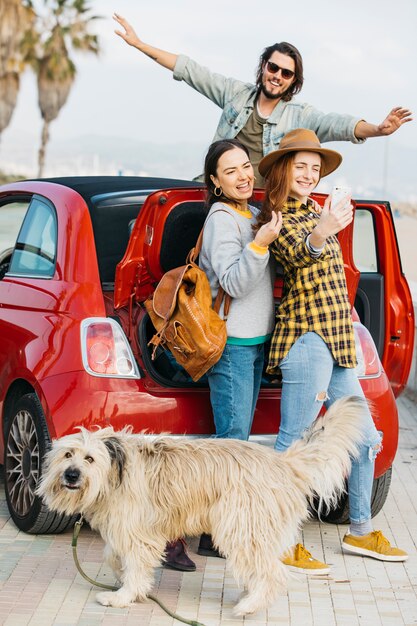 Vrouwen die selfie op smartphone dichtbij autoboomstam en mens nemen die uit auto en hond leunen