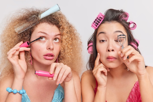 vrouwen make-up staan naast elkaar aanbrengen haarrollers mascara gebruiken wimpers krultang voorbereiden op een speciale gelegenheid er mooi uit willen zien.