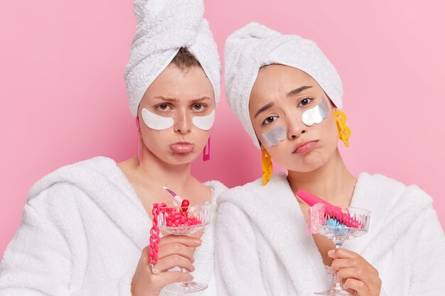 vrouwen passen schoonheidspleisters toe om de huid te hydrateren houden cocktailglazen vol met verschillende accessoires teleurgesteld door iets dragen badjassen handdoek op hoofden geïsoleerd op roze muur