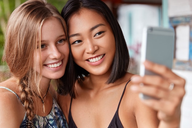 Vrouwen van gemengd ras hebben een waarheidsgetrouwe vriendschap, poseren voor het maken van selfie in een modern café. Multi-etnische jonge vrouwen gebruiken mobiele telefoon om foto's te maken