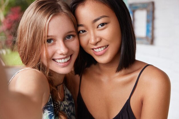 Vrouwen van gemengd ras hebben een waarheidsgetrouwe vriendschap, poseren voor het maken van selfie in een modern café. Multi-etnische jonge vrouwen gebruiken mobiele telefoon om foto's te maken