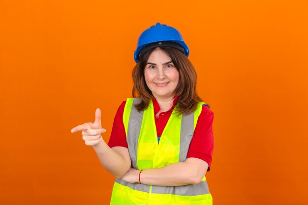 Vrouweningenieur bouwvest dragen en veiligheidshelm die aan iets met glimlach op gezicht richten die zich over geïsoleerde oranje muur bevinden