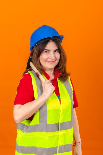 Vrouweningenieur bouwvest dragen en veiligheidshelm die zich met hamer op schouder met glimlach op gezicht over geïsoleerde oranje muur bevinden