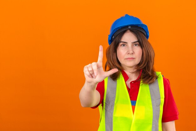 Vrouweningenieur die bouwvest dragen en veiligheidshelm die met omhoog vinger en boze uitdrukking richten die geen gebaar over geïsoleerde oranje muur tonen