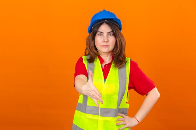 Vrouweningenieur die bouwvest en veiligheidshelm met ernstig gezicht dragen die hand aanbieden die groetgebaar over geïsoleerde oranje muur maken