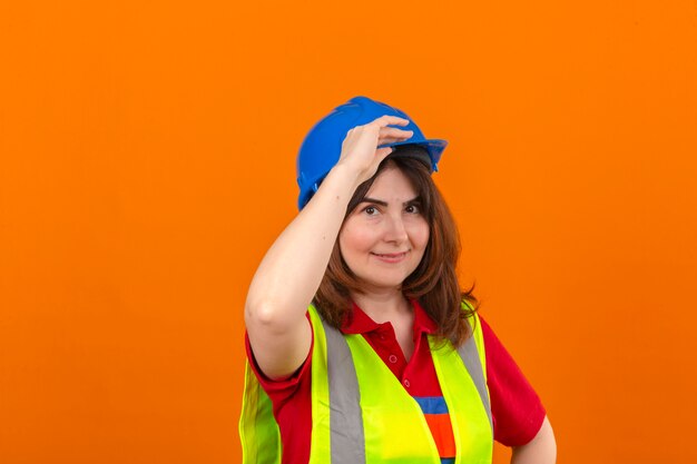 Vrouweningenieur in bouwvest en veiligheidshelm die zeker makend groetgebaar wat betreft helm over geïsoleerde oranje muur kijken kijken