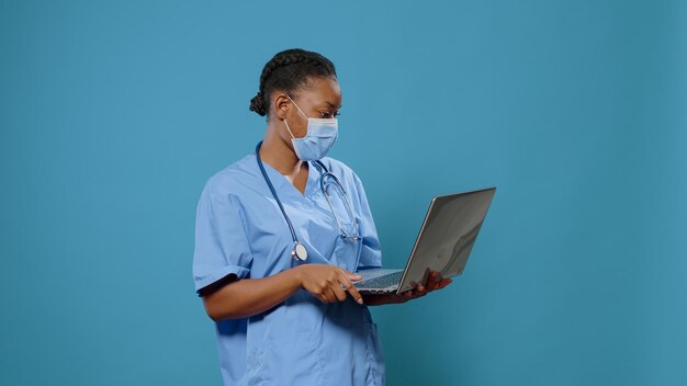 Vrouwenverpleegster die gezichtsmasker draagt en laptop voor camera gebruikt. Medisch assistent in uniform met bescherming tegen coronavirus terwijl hij de computer vasthoudt om aan de gezondheidszorg te werken.