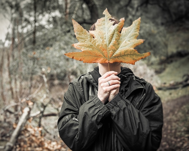 Vrouwtje bedekt haar gezicht met een groot herfstblad terwijl ze midden in een bos staat