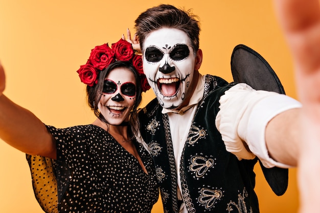 Waanzinnige, grappige jonge man en vrouw nemen selfies en pronken met hun skeletmake-up. Meisje met bloemen op haar hoofd en haar vriend hebben plezier