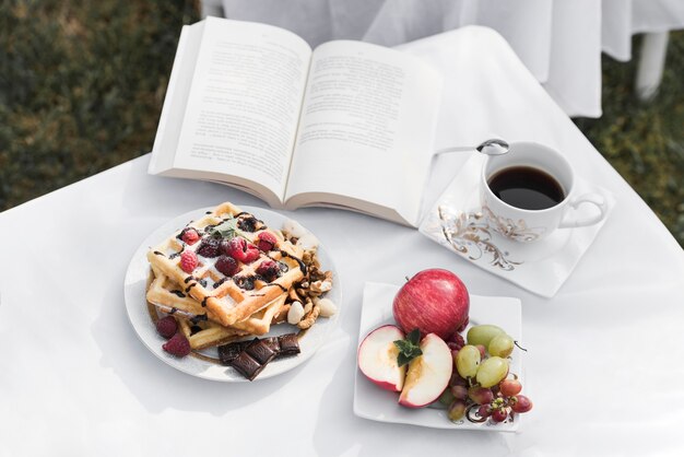 wafels; vruchten; koffiekopje en een open boek op witte tafel