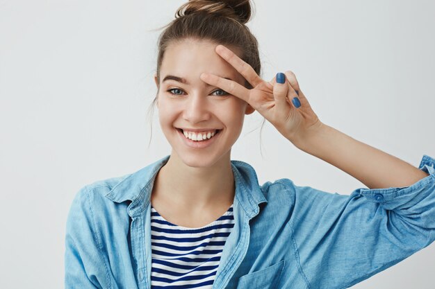 Waist-up studio shot zorgeloos extatisch aantrekkelijke jonge europese vrouw die positief witte tanden glimlacht, vrede toont gelukteken gedrukt hand voorhoofd, gelukkig vrolijk gevoel