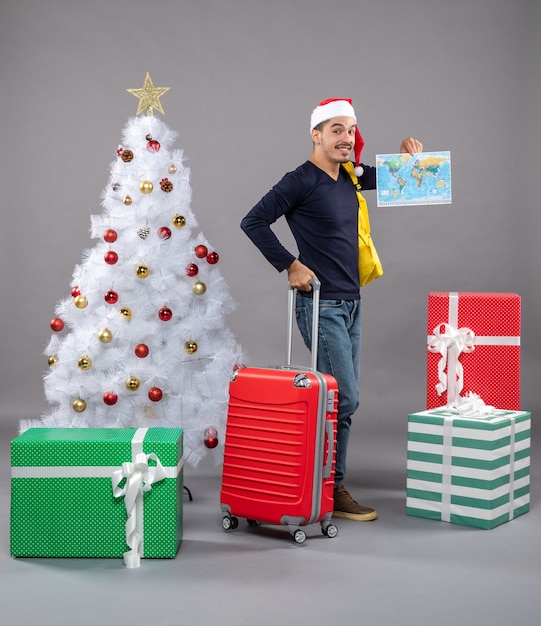 wandelende jonge man met gele rugzak met kaart in de buurt van de kerstboom en presenteert op grijs