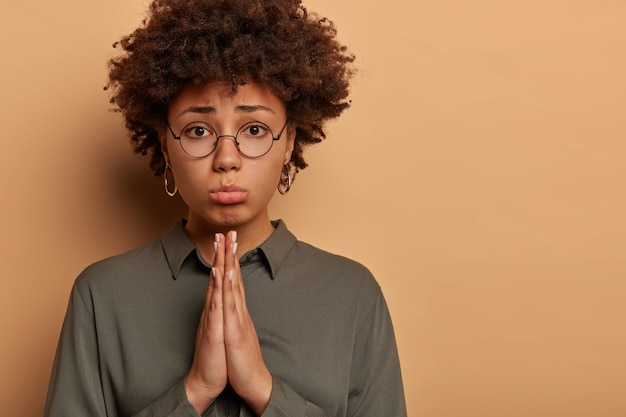 Wanhopige mooie vrouw kijkt met smekend smekend gezicht, vraagt om aplogise, houdt de handpalmen tegen elkaar gedrukt, heeft ogen vol hoop, pakt de onderlip