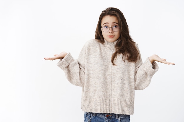 Wat ik fout heb gedaan. Domme en geen idee schattige jonge vrouwelijke student in bril en trui die schouder schouderophalend maakt met handen zijwaarts en onschuldige blik als onbewust en verward over grijze muur.